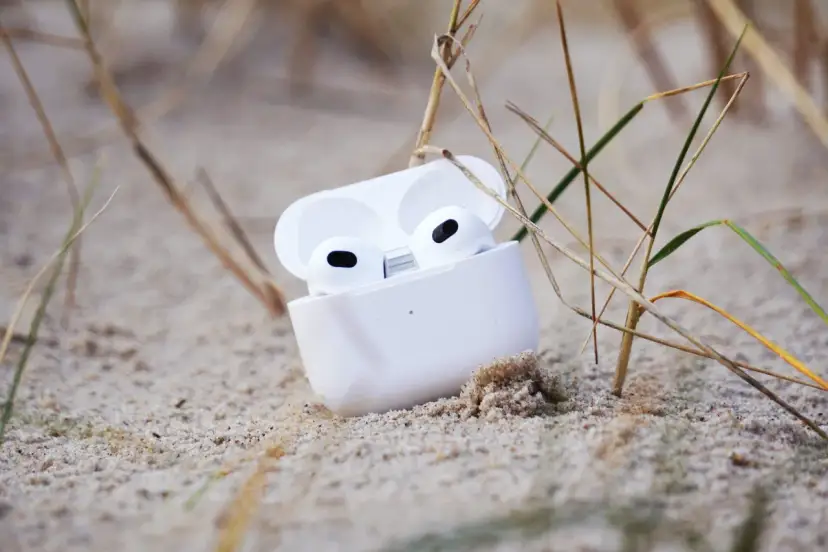 Weiße AirPods im geöffneten Ladecase liegen im Sand, umgeben von Gräsern.