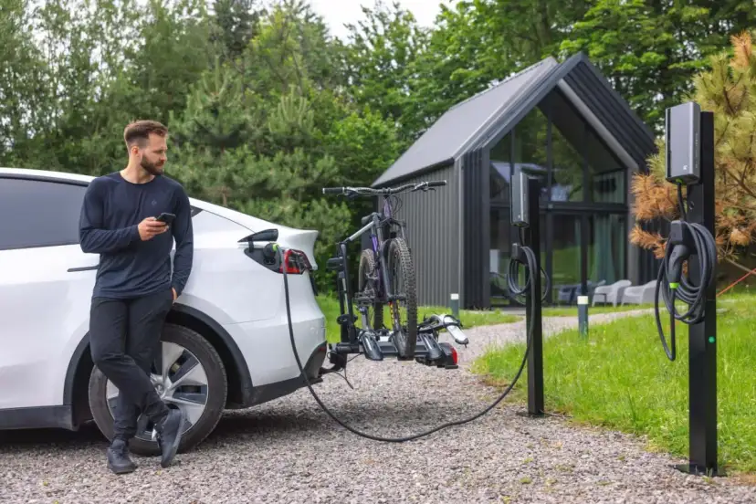Mężczyzna z telefonem przy białym samochodzie elektrycznym, który ładuje się obok stojaka z rowerami. Obok stoją dwie ładowarki do samochodów elektrycznych.