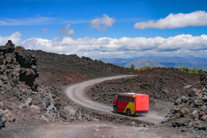Czerwono-żółty autobus turystyczny jedzie krętą drogą przez wulkaniczne krajobrazy Etny.