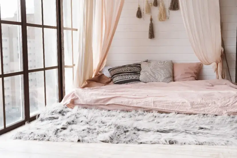 Przytulna sypialnia z łóżkiem i baldachimem DIY, ozdobionym frędzlami. Miękki dywan i duże okno dodają uroku.