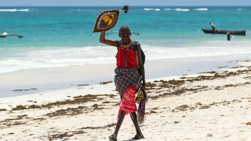 Mężczyzna w tradycyjnym stroju na plaży w Kenii, trzymający ozdobną tarczę. Może to symbolizować bogactwo kulturowe, które jest cenniejsze niż kenijska waluta.