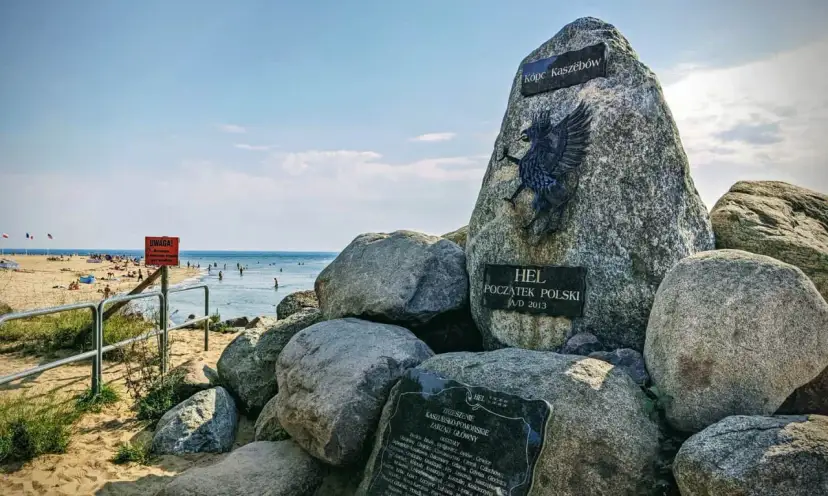Pomnik "Początek Polski" na plaży na cyplu helskim. W tle widać morze i plażowiczów.
