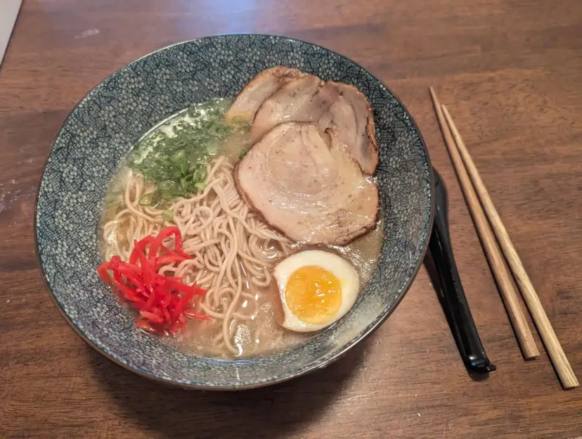 Miseczka z ramen: makaron, plastry mięsa, jajko na pół, zielona cebulka i czerwone przyprawy do ramen. Obok pałeczki.