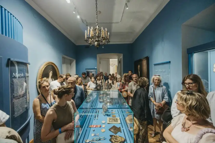 Grupa zwiedzających ogląda wystawę w muzeum. Wśród eksponatów w gablocie widać ozdobne przedmioty, a na ścianach obrazy i tablice informacyjne. Klasyczny wystrój sali z żyrandolem stanowi tło dla tego współczesnego zainteresowania historią.