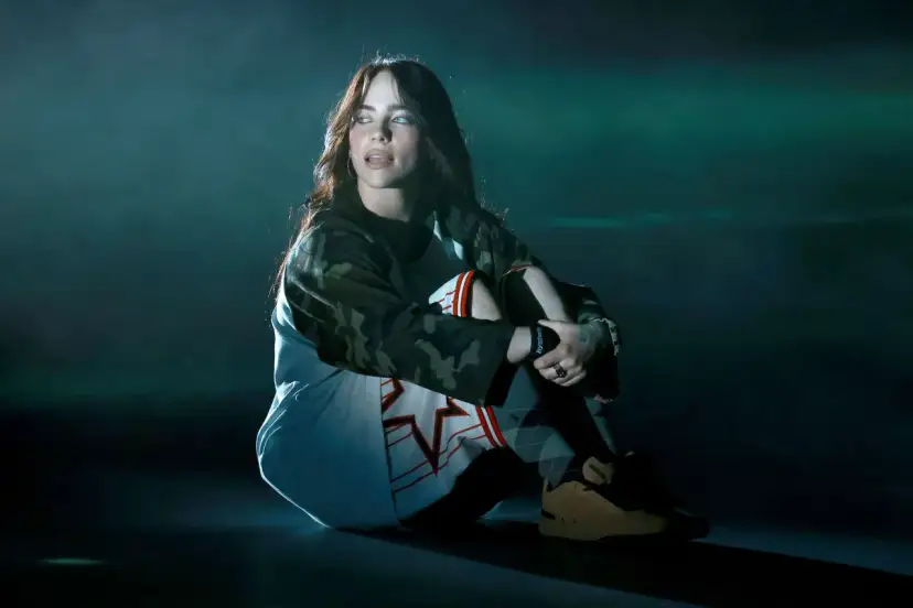 Billie Eilish siedzi na scenie, wpatrzona w dal. Jej stylizacja nawiązuje do estetyki jej albumu.