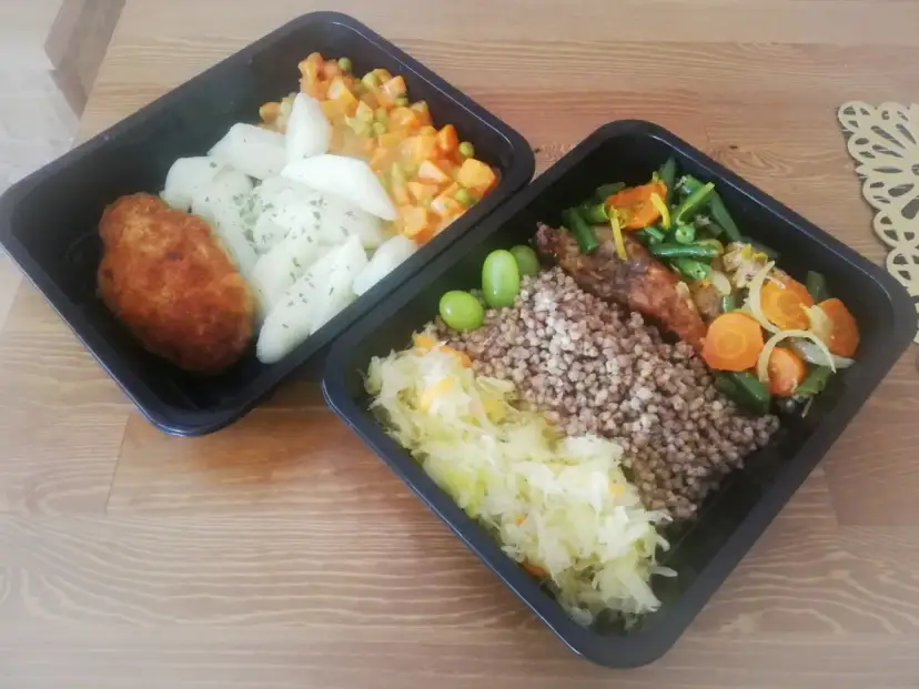 Dwa czarne pojemniki z jedzeniem: kotlet, ziemniaki, warzywa, kasza gryczana, surówka i winogrona.