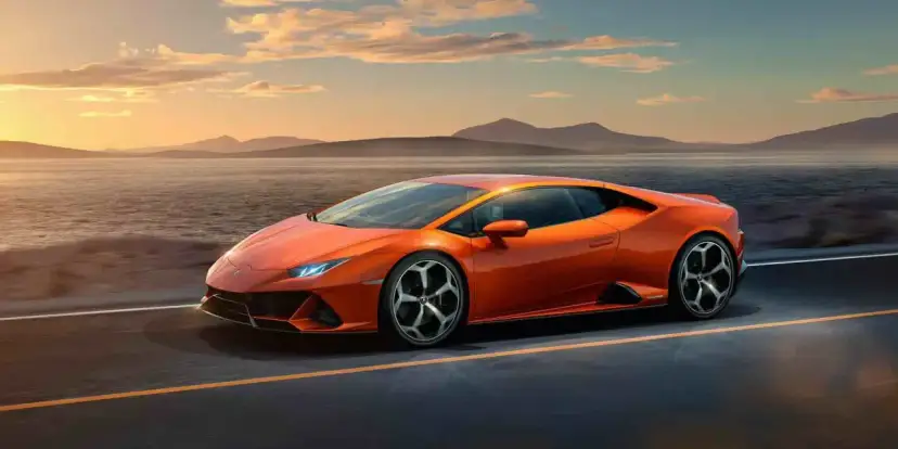 Pomarańczowy Lamborghini Huracan pędzi po pustej drodze nad jeziorem o zachodzie słońca.