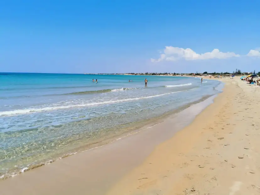 A wide, sandy beach with clear turquoise water and a few people swimming and sunbathing under a bright blue sky.
