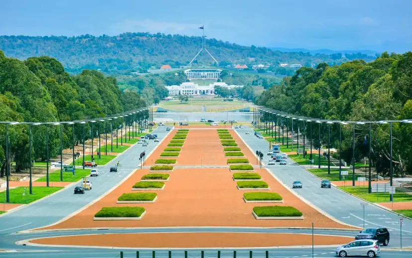 Widok na Parliament House, symbol tego, co jest stolicą Australii. Długa aleja z drzewami i samochodami prowadzi do budynku.