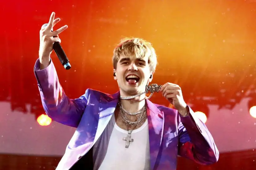 Mata w fioletowej marynarce z naszyjnikiem z napisem "MATA" i mikrofonem na koncercie.