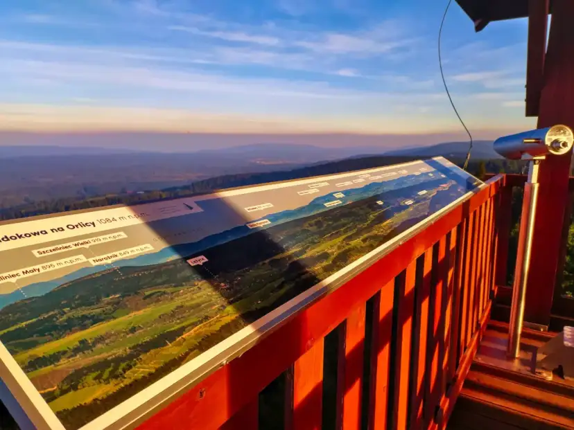Widok z wieży widokowej na Orlicy. Na pierwszym planie panorama z opisem szczytów i dolin. W oddali łagodne wzgórza o zachodzie słońca.