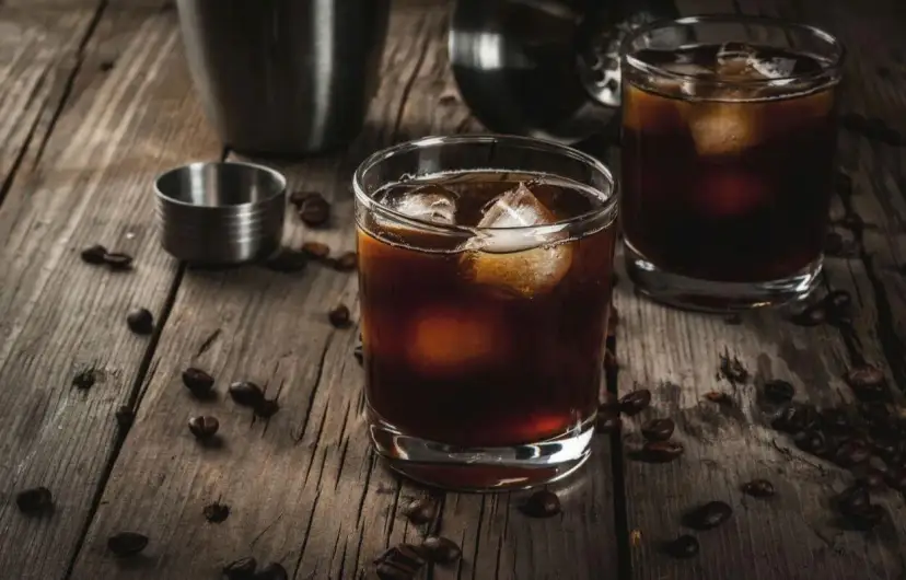 Dwa szklanki z ciemnym napojem i lodem, na drewnianym stole, obok ziaren kawy i shakera.