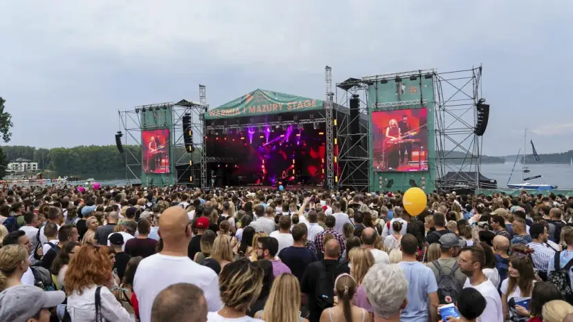 Tłum ludzi na koncercie pod gołym niebem. Na scenie gra zespół, a na ekranach wyświetlany jest green festival line up.