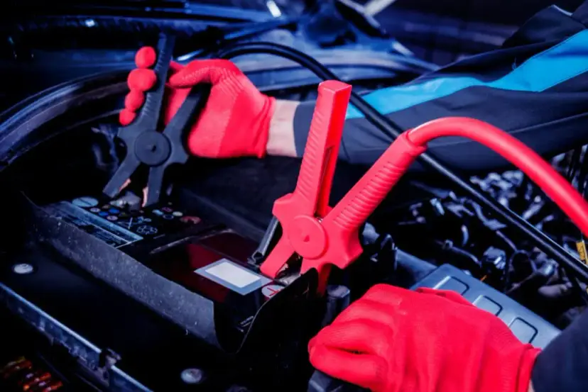 Ręce w czerwonych rękawiczkach podłączają kable prostownika do akumulatora samochodowego.