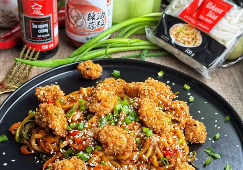 Pyszny makaron udon z kurczakiem w panierce, posypany sezamem i szczypiorkiem. Obok sosy i opakowanie makaronu.