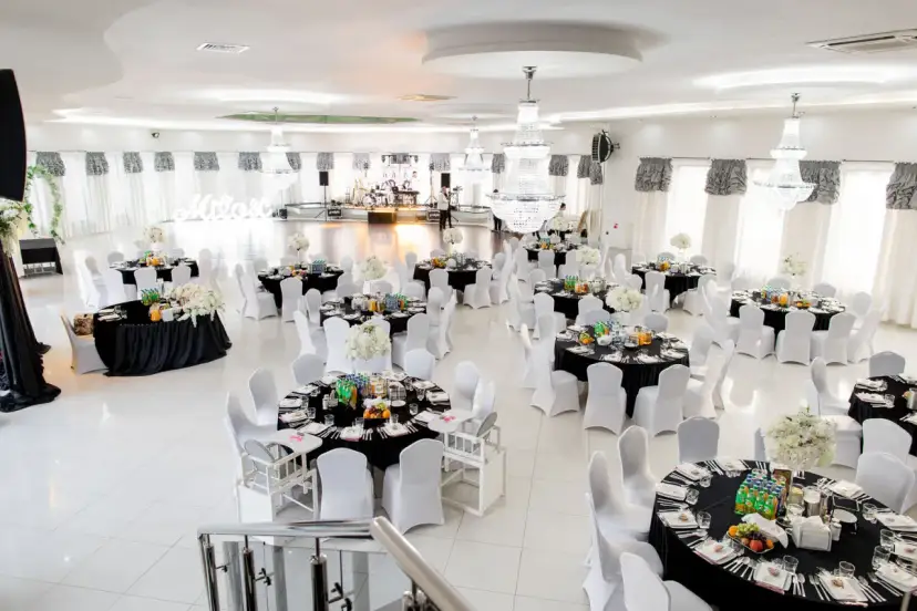 Elegancka sala weselna z czarnymi stołami, białymi krzesłami i kryształowymi żyrandolami.