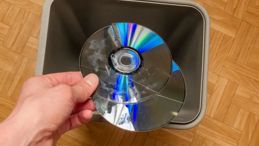 Dłoń trzyma pękniętą płytę CD/DVD nad szarym koszem na śmieci.