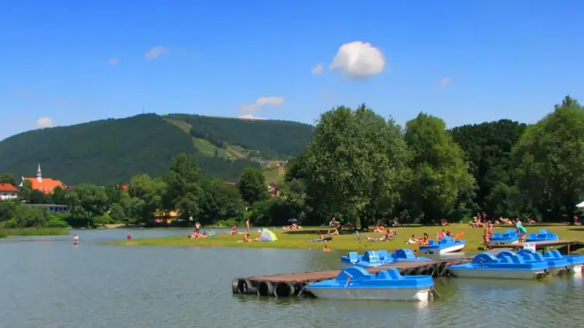 Słoneczny dzień nad jeziorem w Międzybrodziu Żywieckim. Ludzie odpoczywają na plaży, a przy brzegu cumują niebieskie rowerki wodne.