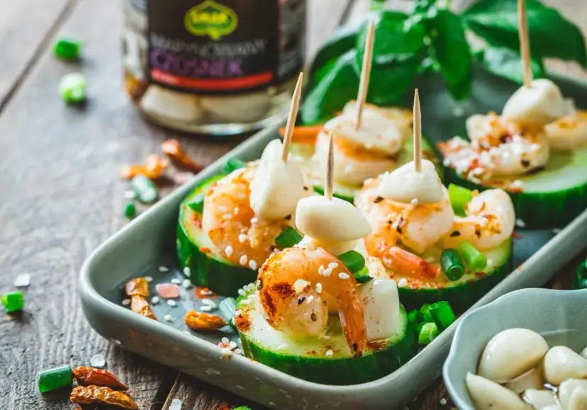 Pyszne koreczki z krewetkami na plastrach ogórka, ozdobione sezamem i szczypiorkiem. Obok słoik marynowanego czosnku.