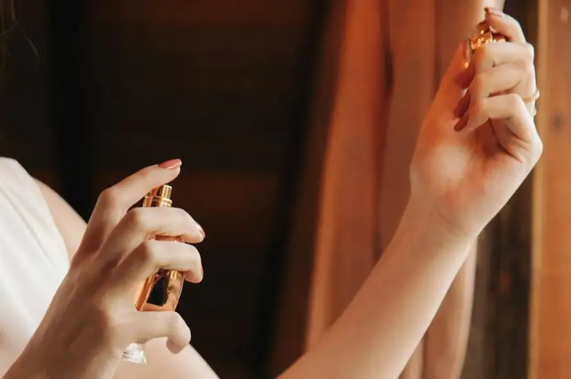 Dłoń kobiety trzymająca złoty flakonik perfum i aplikująca je na skórę.