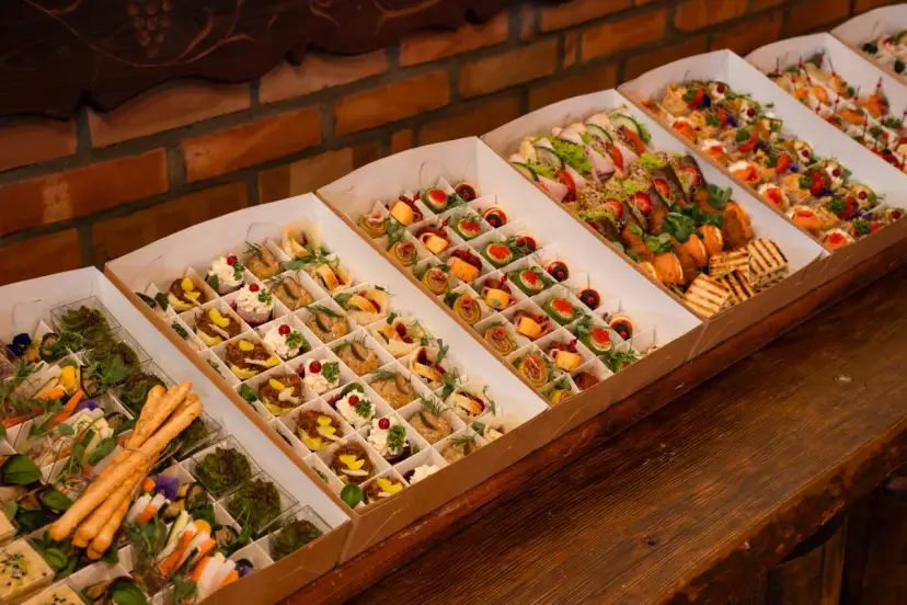 Elegancki catering do domu: pięknie ułożone przekąski w kartonowych pudełkach, od mini kanapek po rolki z łososiem i warzywami.