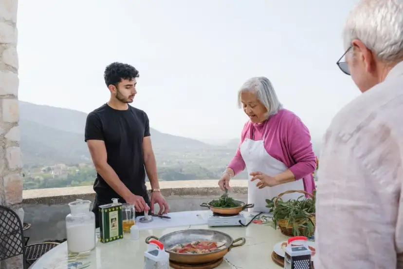 An older woman in an apron cooks greens while a young man and an older man stand nearby on a terrace with a scenic view.