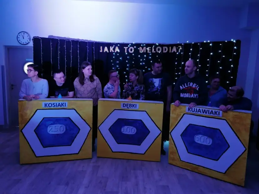 Uczestnicy zabawy "Jaka to melodia?" stoją przy planszach z nazwami drużyn: Kosiaki, Dębki, Kujawiaki.
