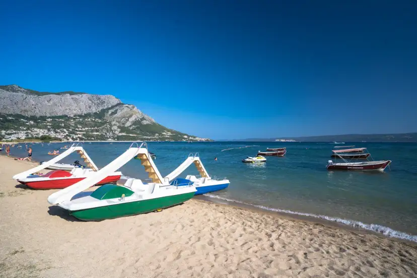 Plaża z łódkami i zjeżdżalniami, idealna na wakacje w camping Galeb. W tle góry i turkusowe morze.