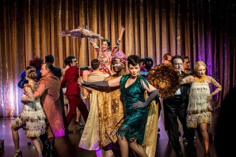 Tancerze w barwnych strojach, jak żołnierz królowej Madagaskaru, na scenie z błyszczącą kurtyną.