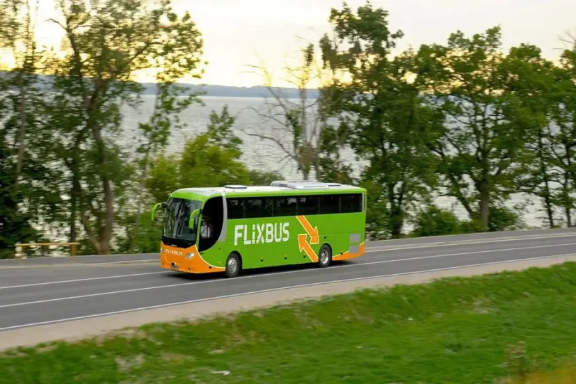 Zielony autobus Flixbus jedzie drogą przy jeziorze otoczonym drzewami.