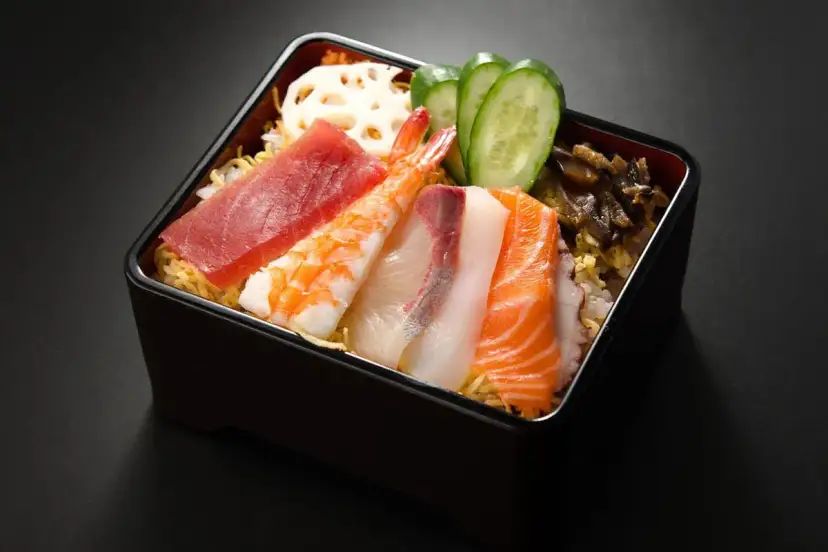 Chirashi zushi: kolorowe plastry ryb (tuńczyk, łosoś, biała ryba), krewetka, plasterki ogórka i lotosu na ryżu.