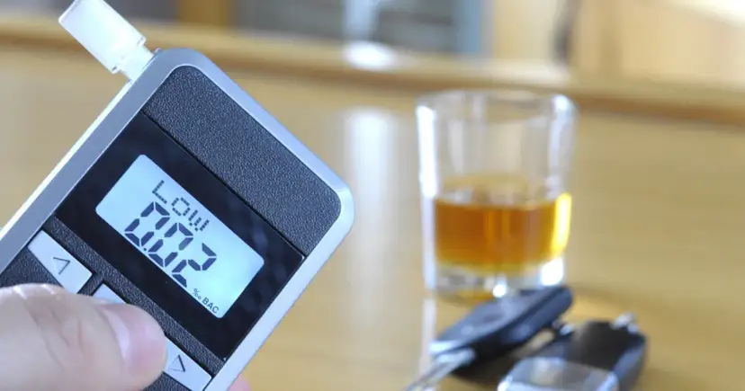 Alkomat z wyświetlaczem pokazującym 0.02% BAC, obok kieliszek z alkoholem i kluczyki samochodowe.