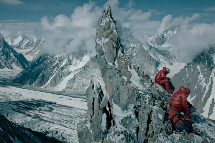 Dwóch alpinistów w czerwonych kurtkach odpoczywa na skalistym szczycie, podziwiając lodowcową dolinę i ośnieżone góry.