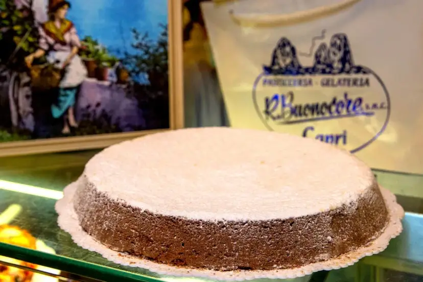 A Torta Caprese dusted with powdered sugar sits on a glass counter, with a painting and shop logo in the background.