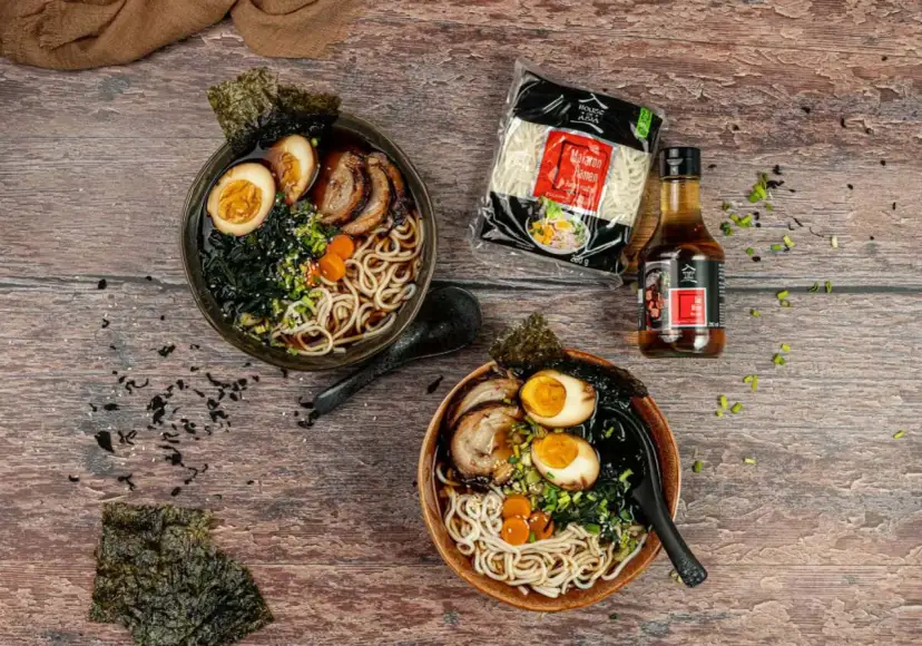 Dwa miski aromatycznego ramenu z boczkiem, jajkiem i nori. Obok opakowanie makaronu i sosu.