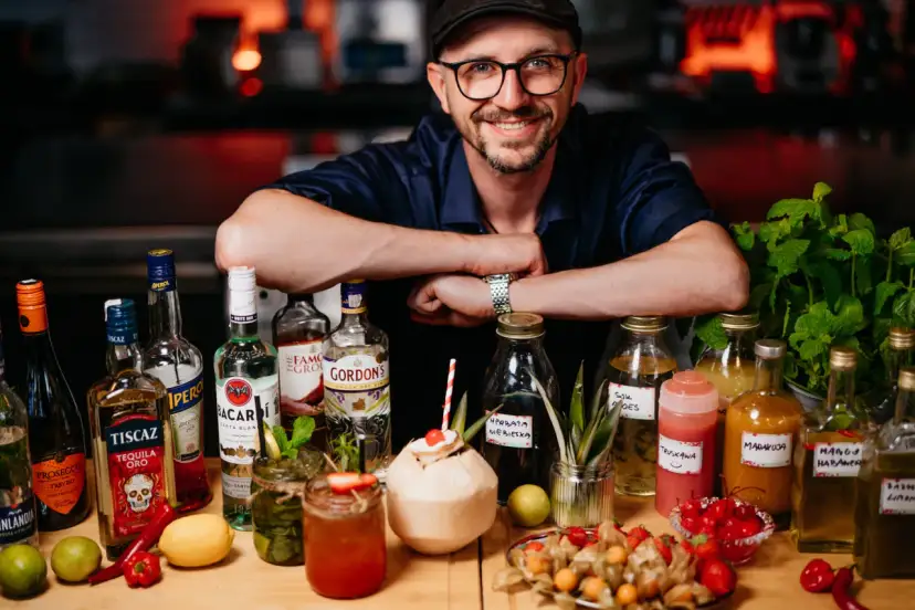 Uśmiechnięty mężczyzna w czapce i okularach za ladą pełną alkoholi i owoców. To przyszłość: drink bar zamiast barmana.
